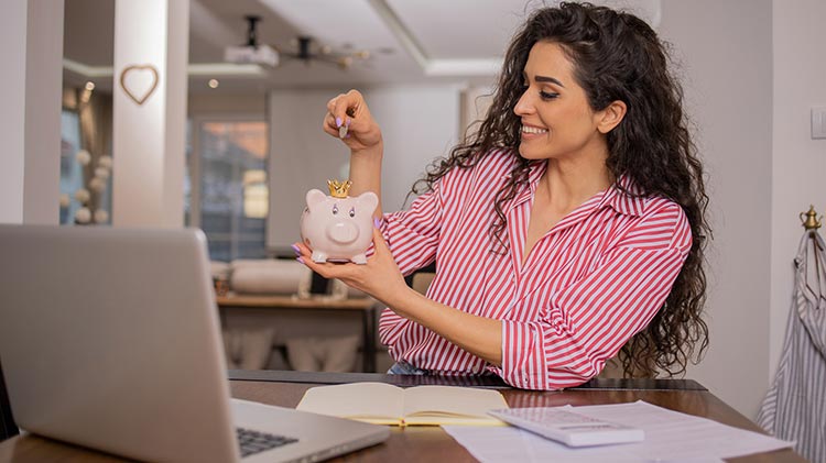 Woman with bills, laptop and calculator in front of her and adding money in a piggy bank.