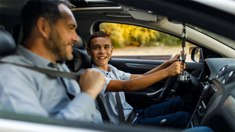 A father putting on his seat belt in his teen son’s first car.