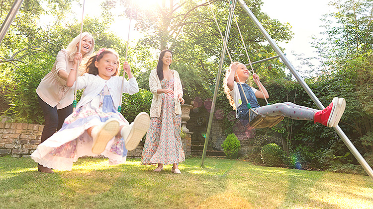 Two women pushing young girls on a backyard playground.