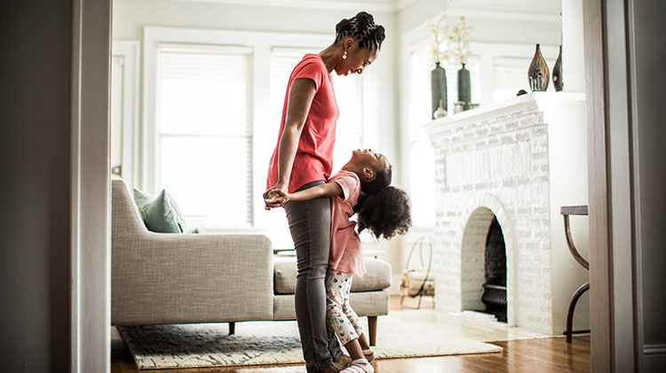 Little girl smiling and looking up at her mother while standing on her mother’s feet in their living room.